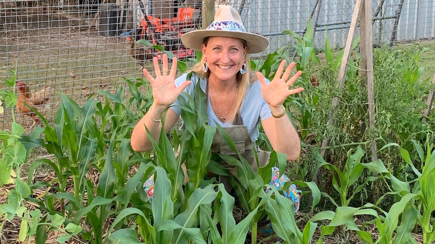 A woman wearing a sunhat squats in among rows of plants in a vege plants, she is grinning and her hands are raised and open.