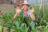 A woman wearing a sunhat squats in among rows of plants in a vege plants, she is grinning and her hands are raised and open.
