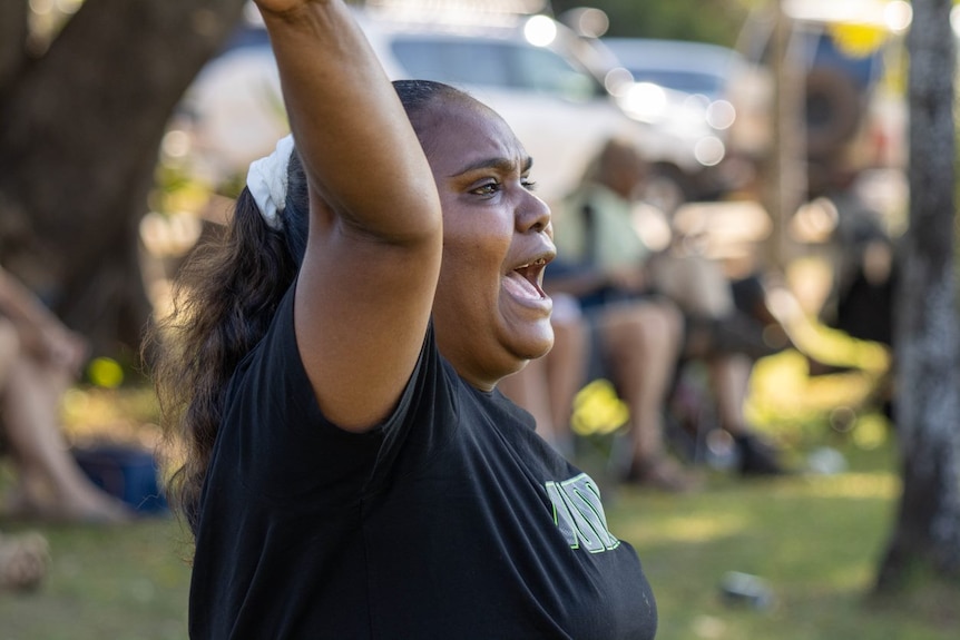 A woman with her arms up excitedly yelling from the sideline of a footy game. 