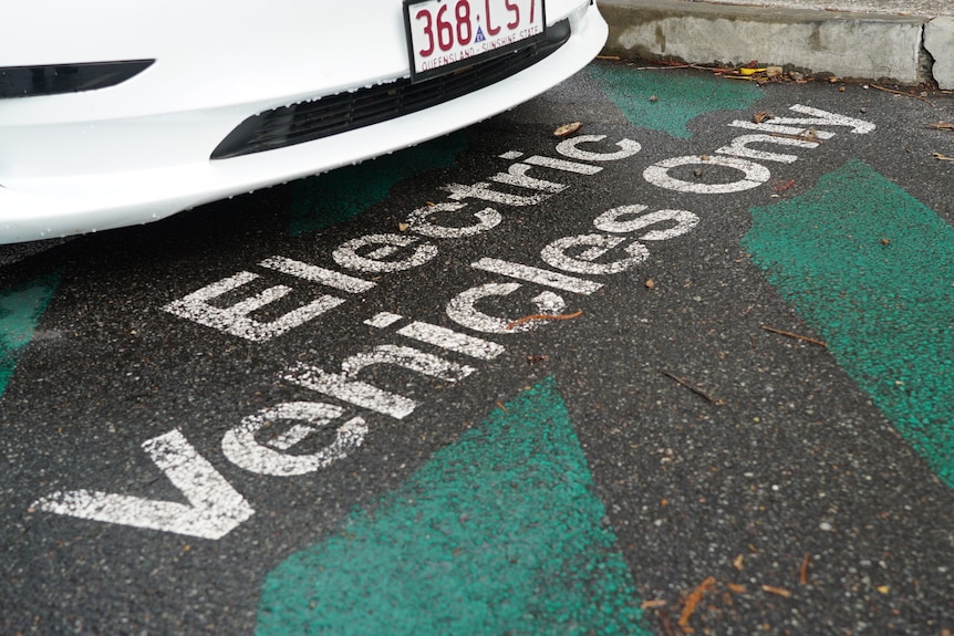 A white car is stopped in a Brisbane charging station carpark, white 'electric vehicles only' text is written over green lines