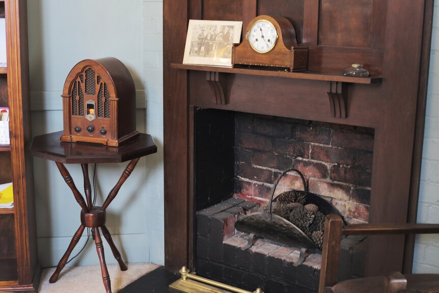 Pine comes sit in a brick fireplace. On the mantel is a wooden clock. On a wooden table to the left is an old wooden radio.
