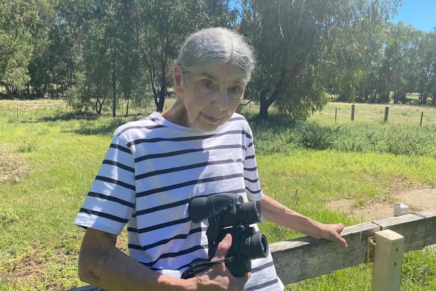 A woman holding binoculars while leaning against a fence in her backyard