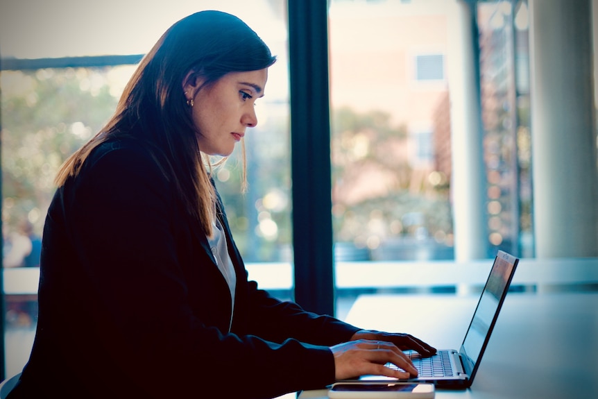 The side profile of a woman with dark hair and a dark blazer working on a laptop.