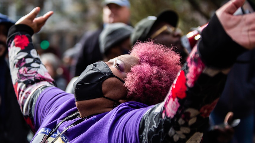 A woman with pink hair looks up to the sky, arms outstretched