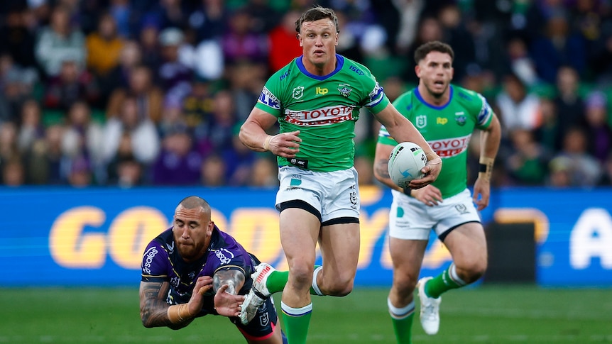 A Canberra Raiders NRL player holds the ball in his left hand as he beats a Melbourne Storm defender.