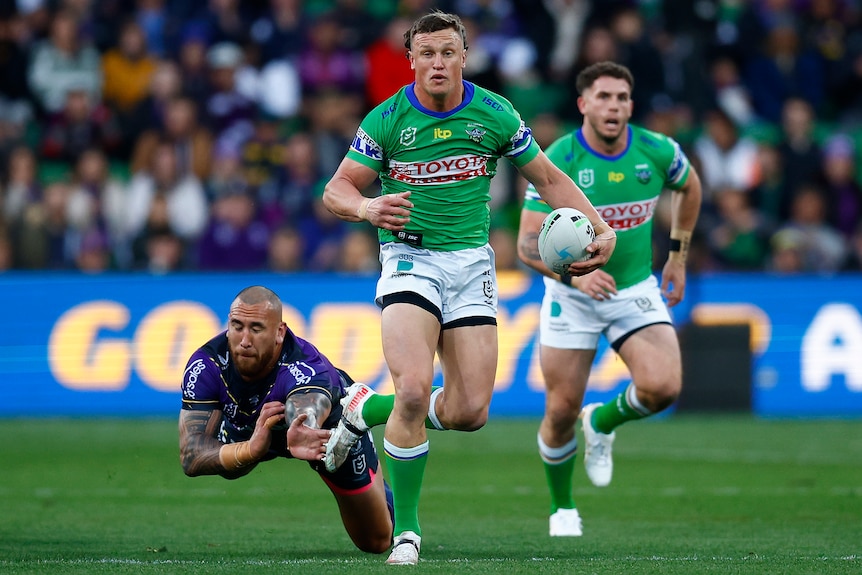 A Canberra Raiders NRL player holds the ball in his left hand as he beats a Melbourne Storm defender.