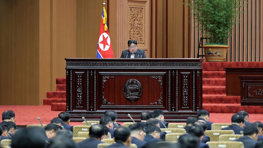Kim Jong Un sits at a large desk in fromt of a room of men