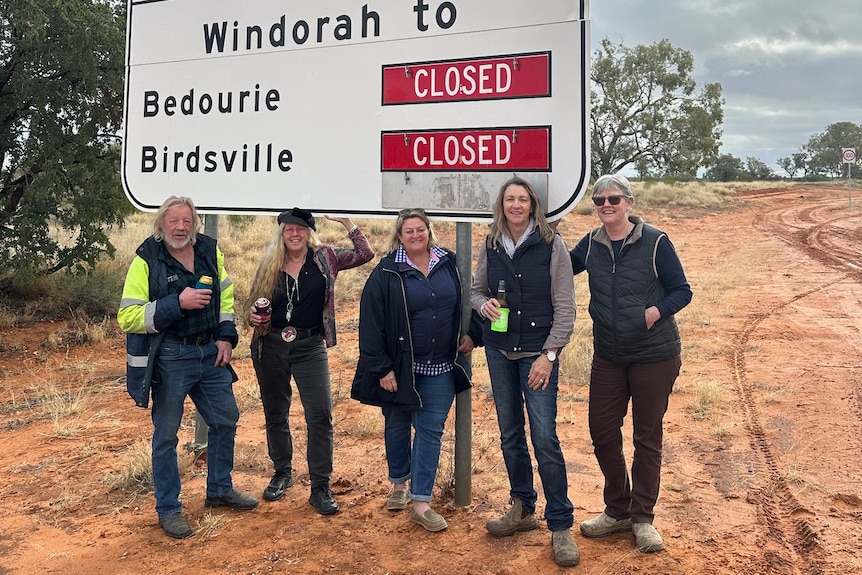 Five people stand in front of a sign that says the road to Birdsville and Bedourie are closed.