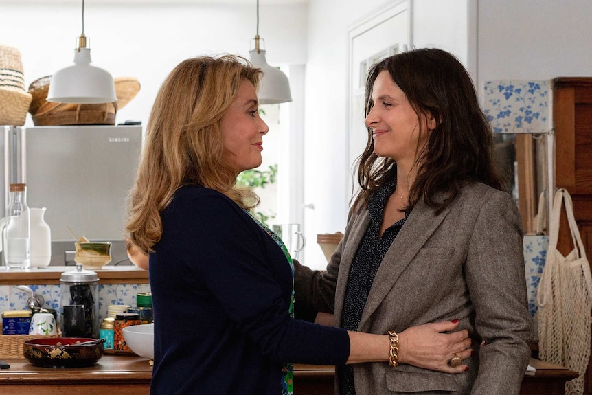 Actors Catherine Deneuve and Juliette Binoche embrace in a kitchen