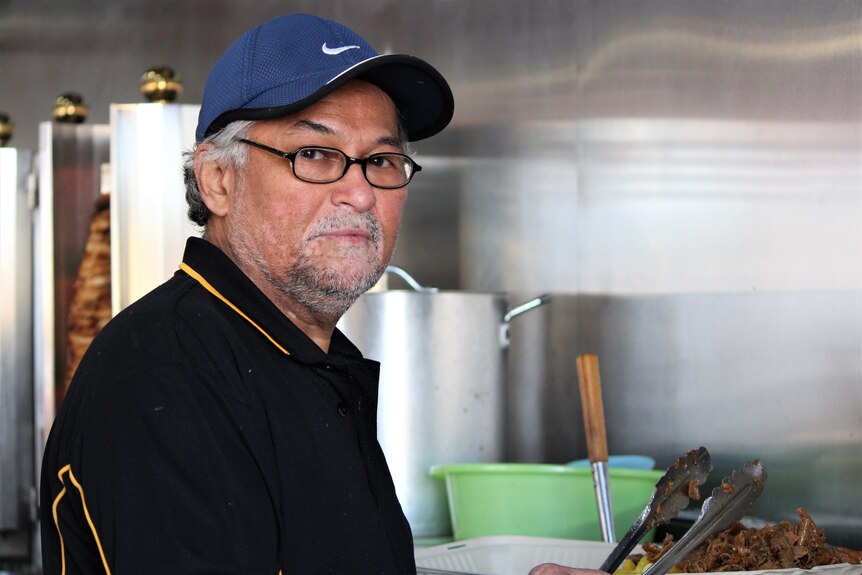 A man with grey hair and a grey beard wears glasses and a blue hat, standing in a kitchen of a kebab shop. 