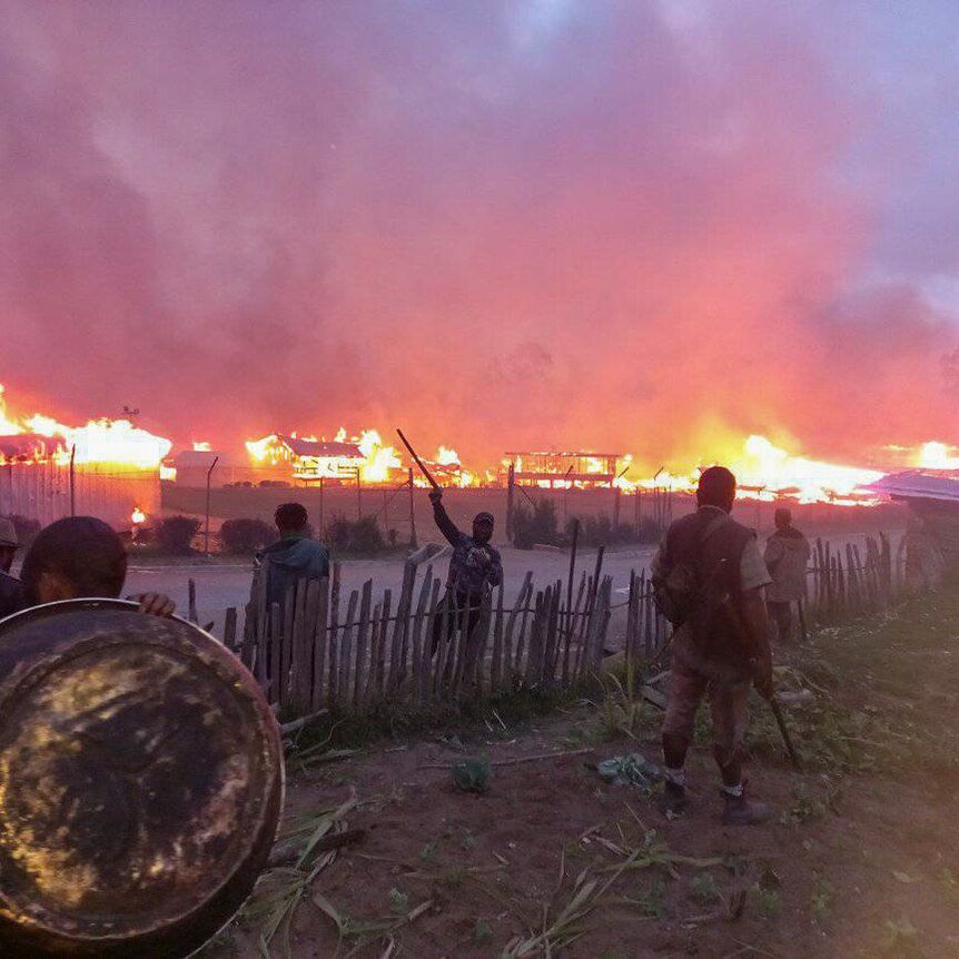 A group of men wielding sticks stand in front of a burning village 