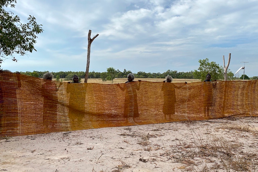 Six First Nations women in the remote Northern Territory hold up a woven 'fish fence', a large piece of fabric.