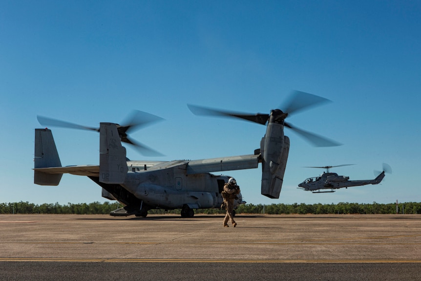 A US Marine Corps MV22 Osprey, which looks like a winged helicopter, sits at idle while a more traditional helicopter takes off.