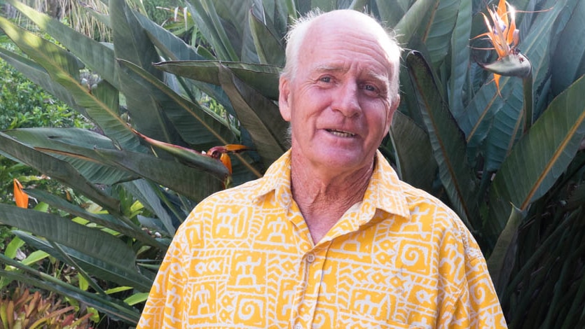 An older man in yellow shirt standing in front of a bird of paradise plant.