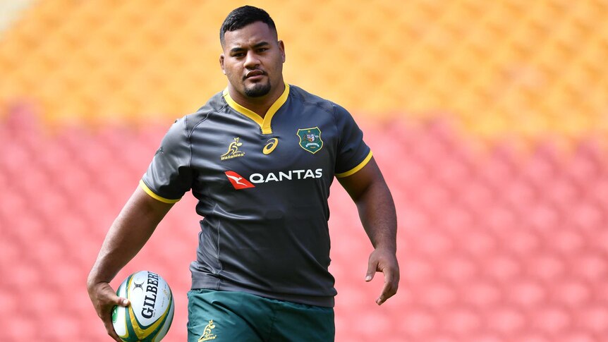 A male rugby union player, wearing a Wallabies training jersey, carries a rugby ball in his right hand as he walks.