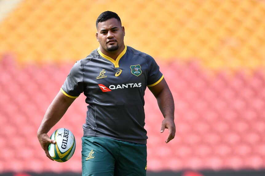 A male rugby union player, wearing a Wallabies training jersey, carries a rugby ball in his right hand as he walks.
