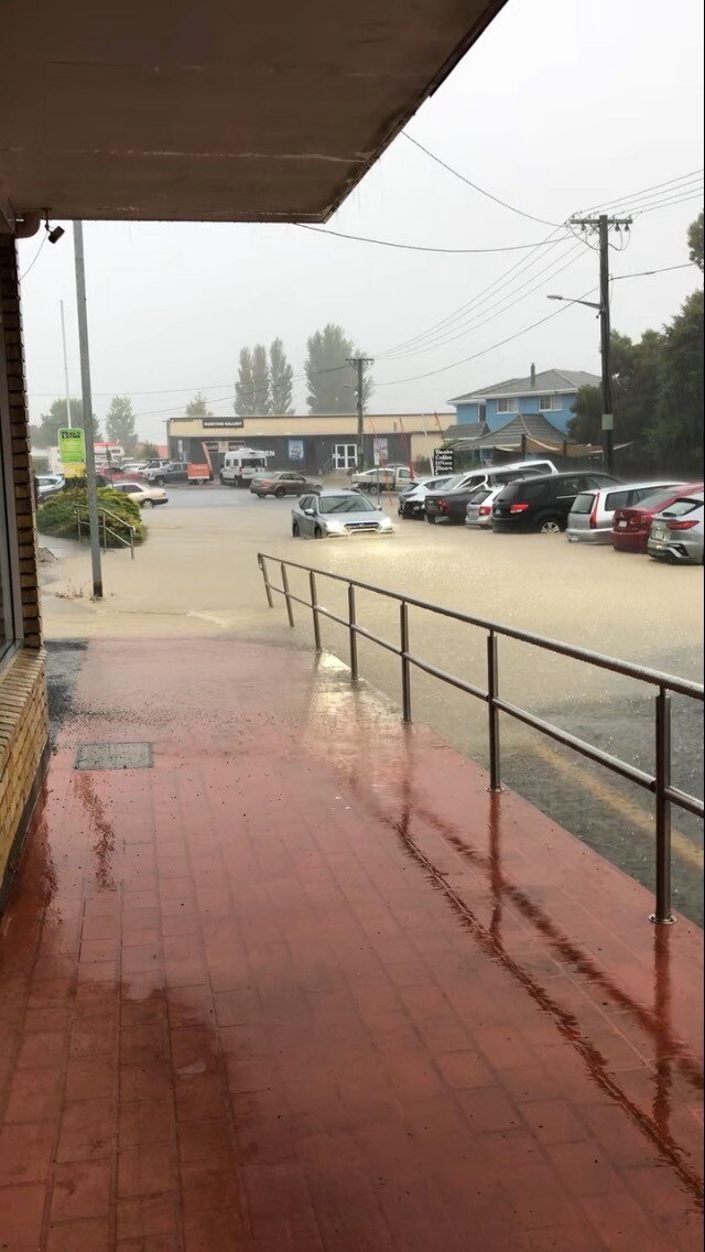 A flooded street in a town showing parked cars sitting in water and a car driving through floodwater.