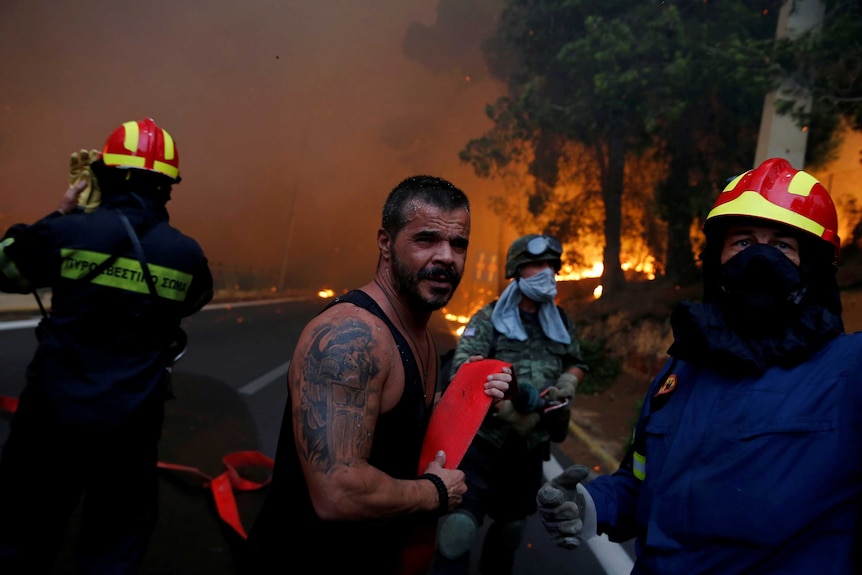 Firefighters wearing helmets and a man in a singlet carry an orange fire hose as flames burn in the background