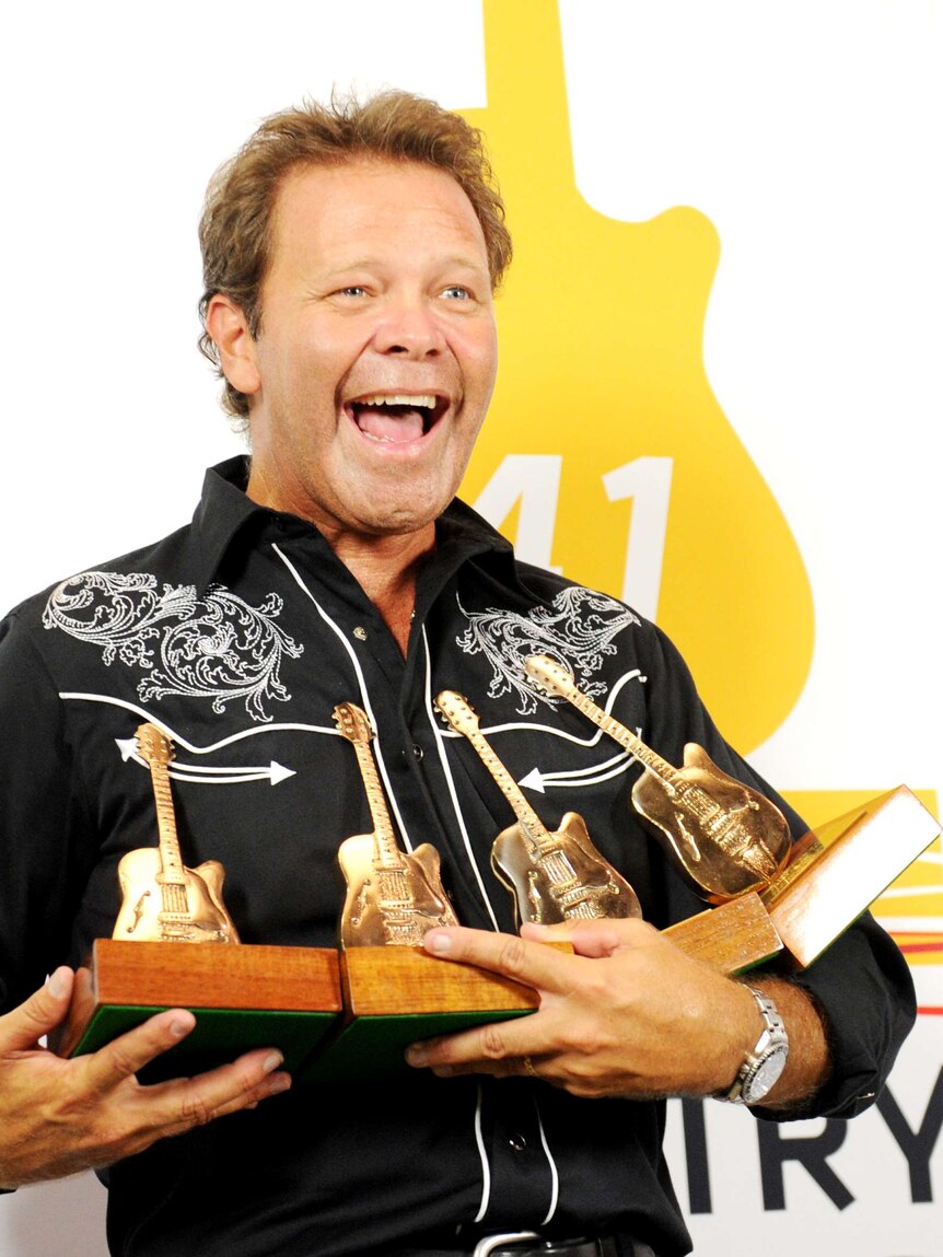 Country musician Troy Cassar-Daley smiles and holds golden guitar awards