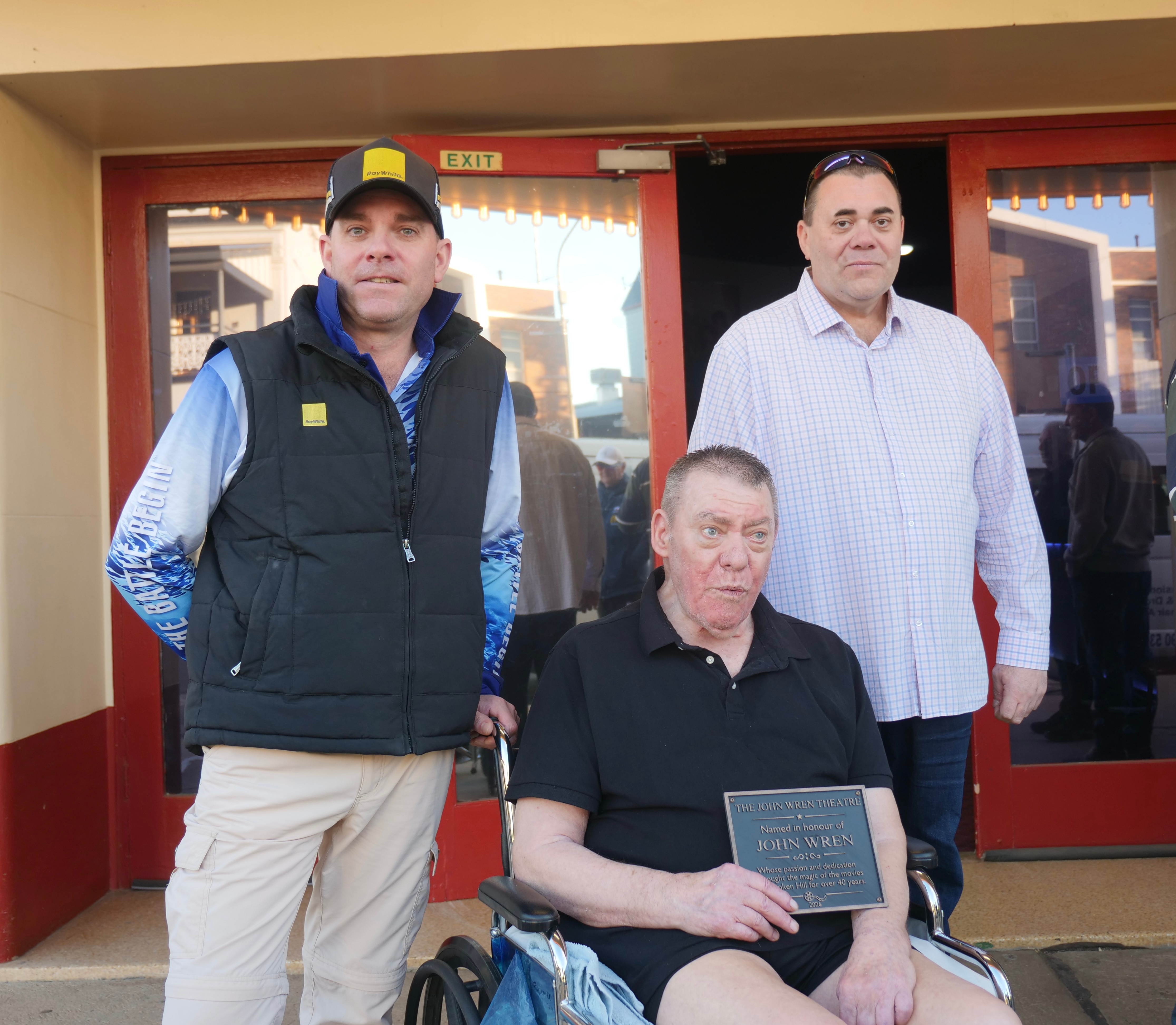 Dos hombres están a cada lado de un anciano en silla de ruedas sosteniendo una placa frente a un cine.