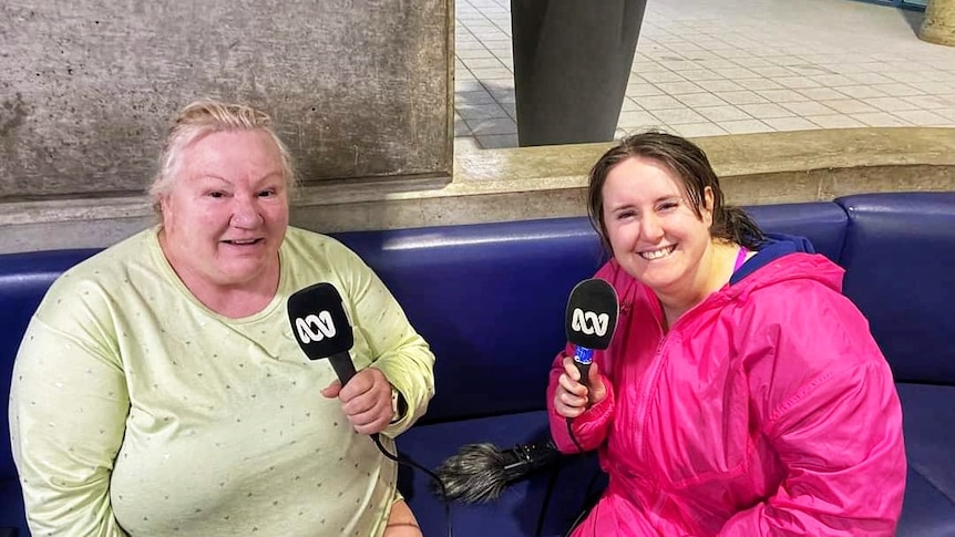 Two smiling women sit at the AIS pool, looking wet, but happy and holding microphones.