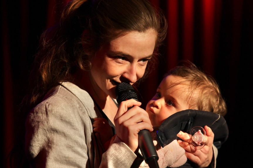 A woman holding a mic on stage while holding a baby.