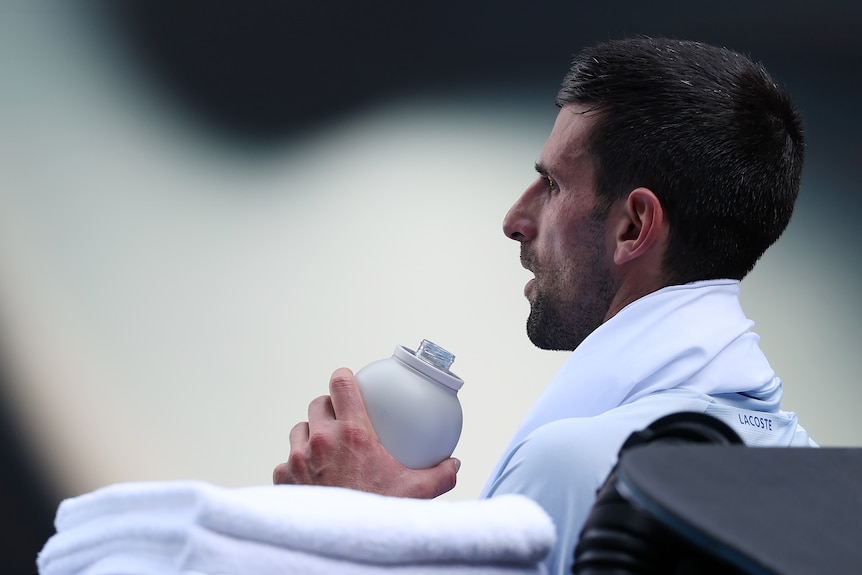 Novak Djokovic, in white shirt, sitting down next to the court, with a white towel over his next, holding a drink bottle 