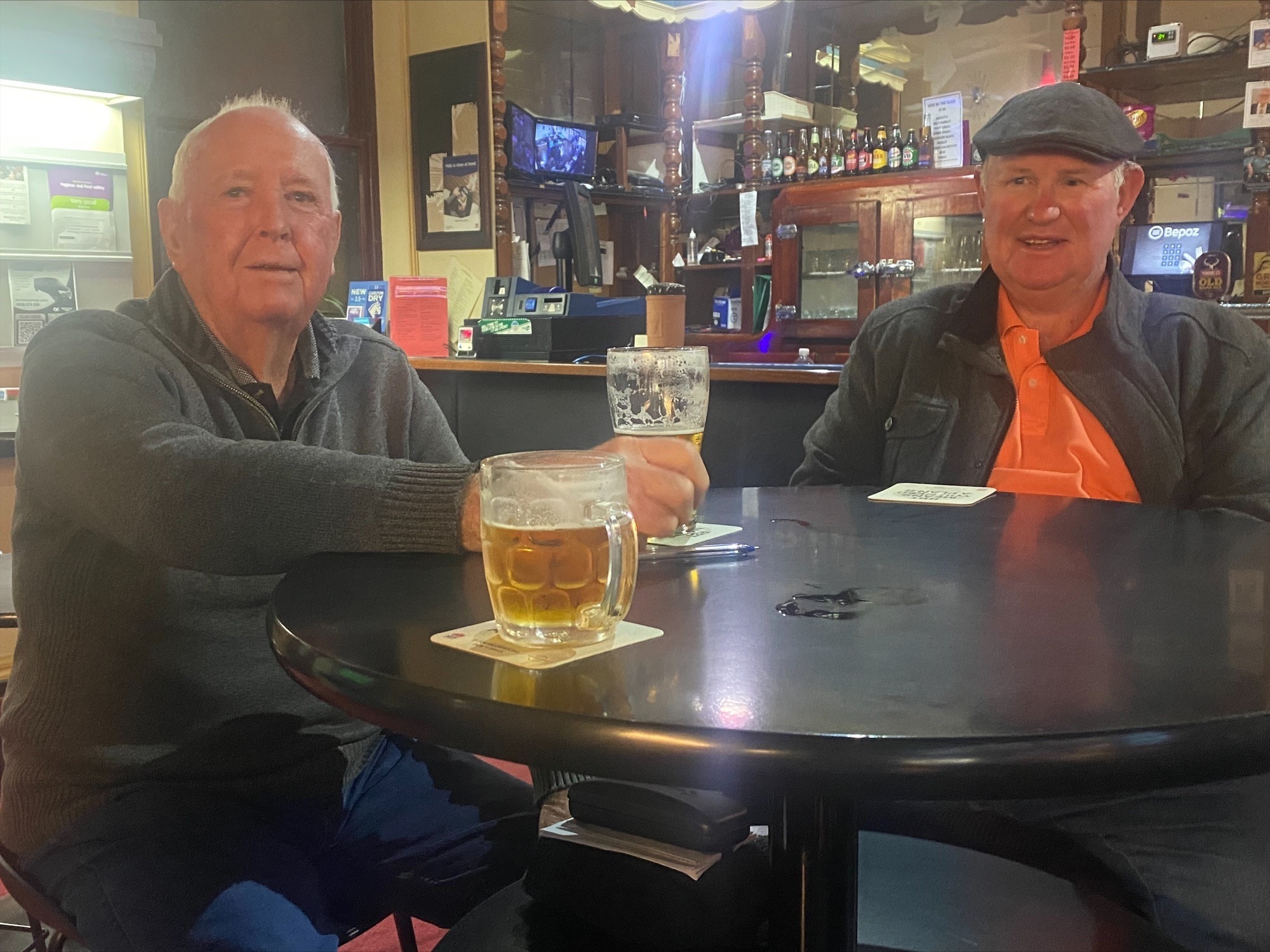 John Kinnear and Michael Baldwin sit at a high top table in a pub with beers.