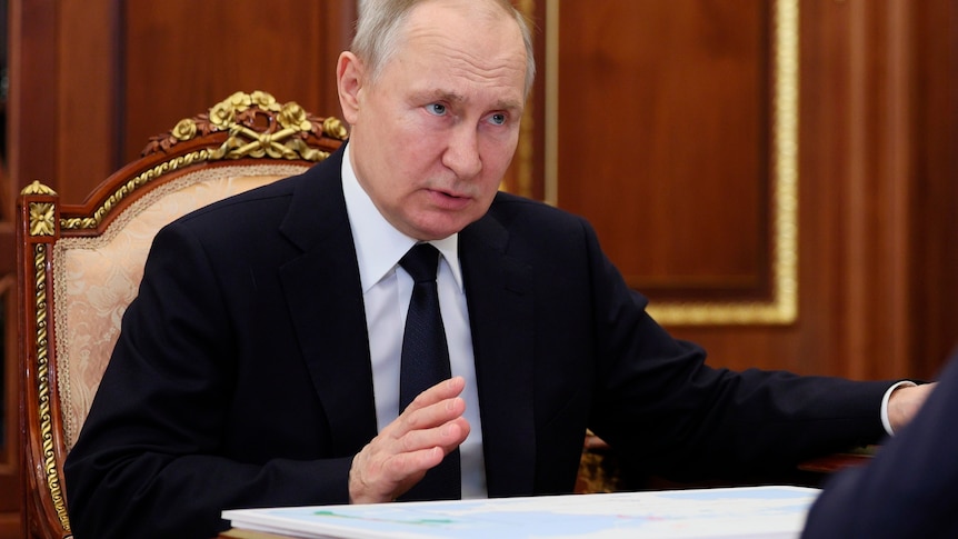 Putin sits on an ornate chair, lifting a hand slightly in gesture as he speaks to someone out of frame