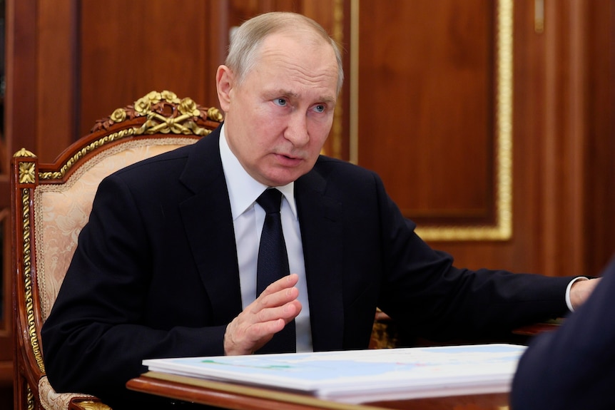Putin sits on an ornate chair, lifting a hand slightly in gesture as he speaks to someone out of frame