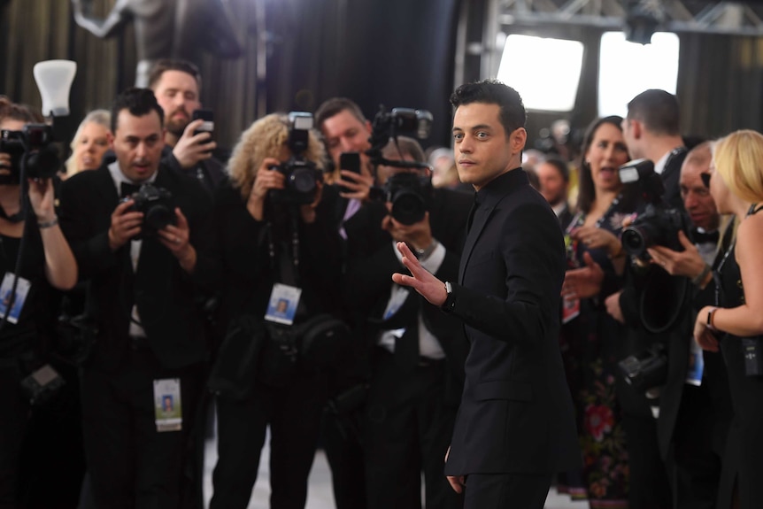 Rami Malek looks to the left wearing black tuxedo as he is surrounded by photographers on red carpet with lights in background.