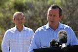Mark McGowan talks at a lecturn with microphones, with Roger Cook in the background.