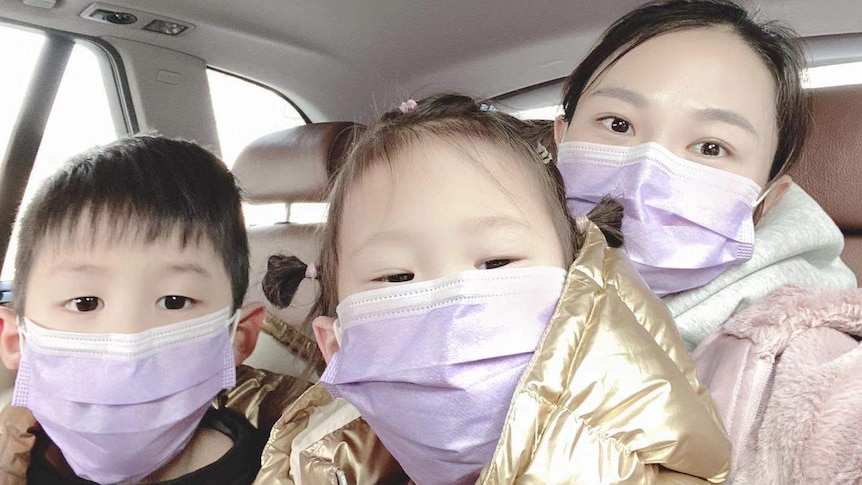 A woman sits in a car with her two children. They are all wearing protective masks.