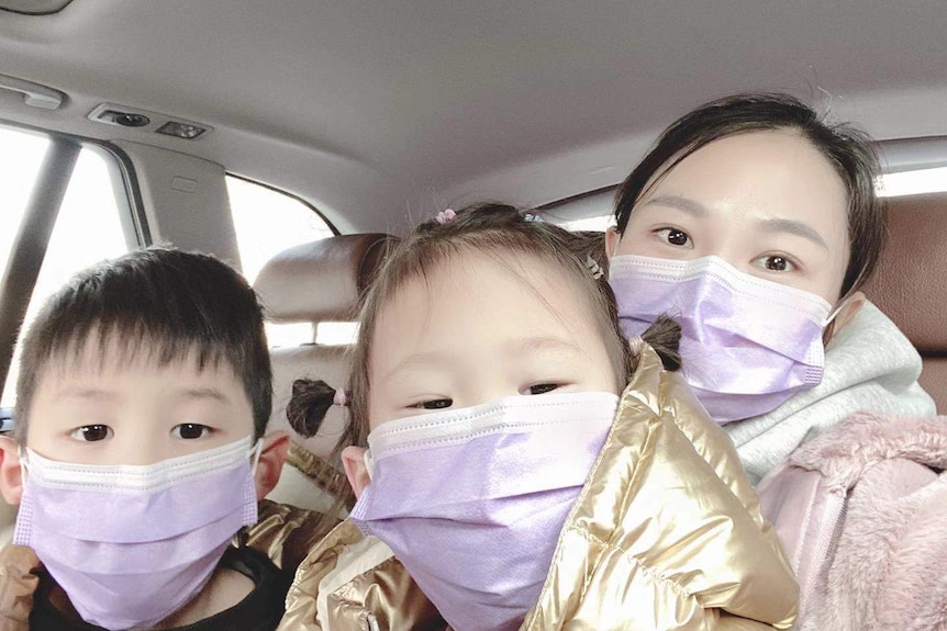 A woman sits in a car with her two children. They are all wearing protective masks.