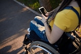A young woman in denim overalls and a yellow T-shirt sits in a wheelchair outdoors, using a smartphone. 