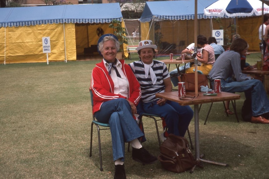 Colour photo of two women sitting under outdoor umbrellas smiling, sitting near a table with cans of coke on it.