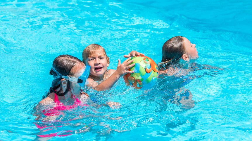 Three children playing in a swimming pool, the boy in the middle is holding a colourful beach ball