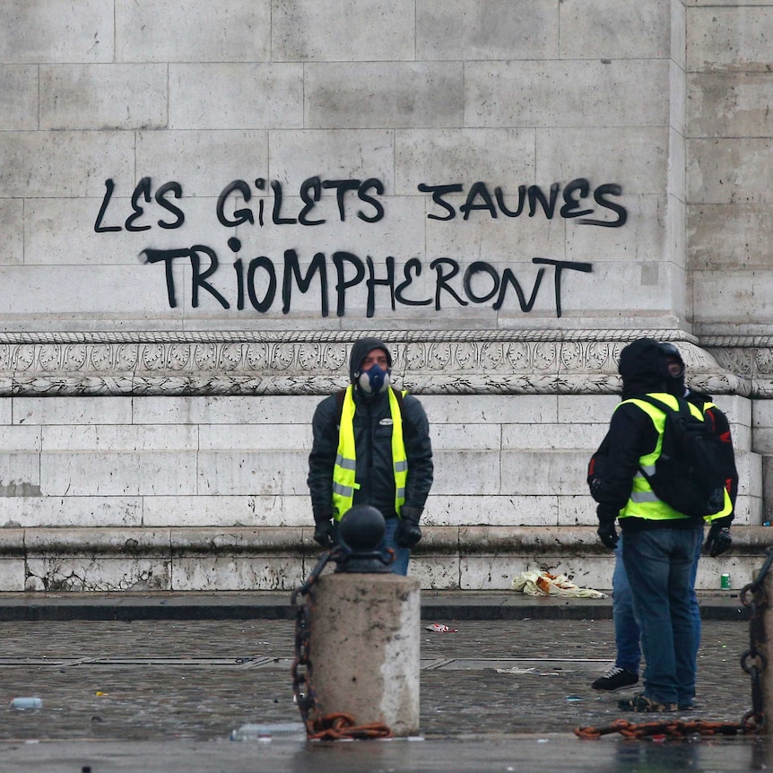 Four protesters wearing bright yellow vests stand outside the graffiti sprayed at the base of the Arc de Triomphe