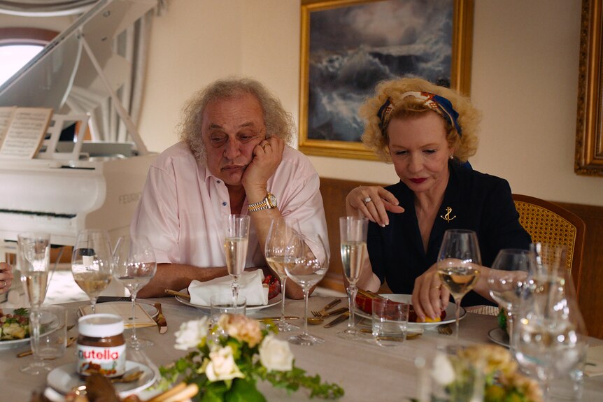 Three wealthy white people - two older and one young - at a table with sparkling wine on a boat, white grand piano in background