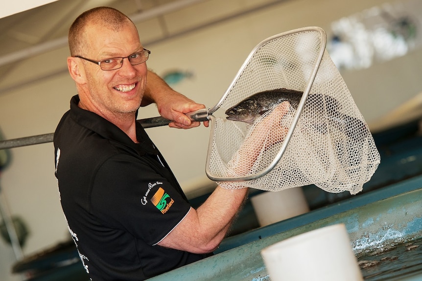 A smiling man holds up a fish inside a net that he is holding above a tank.