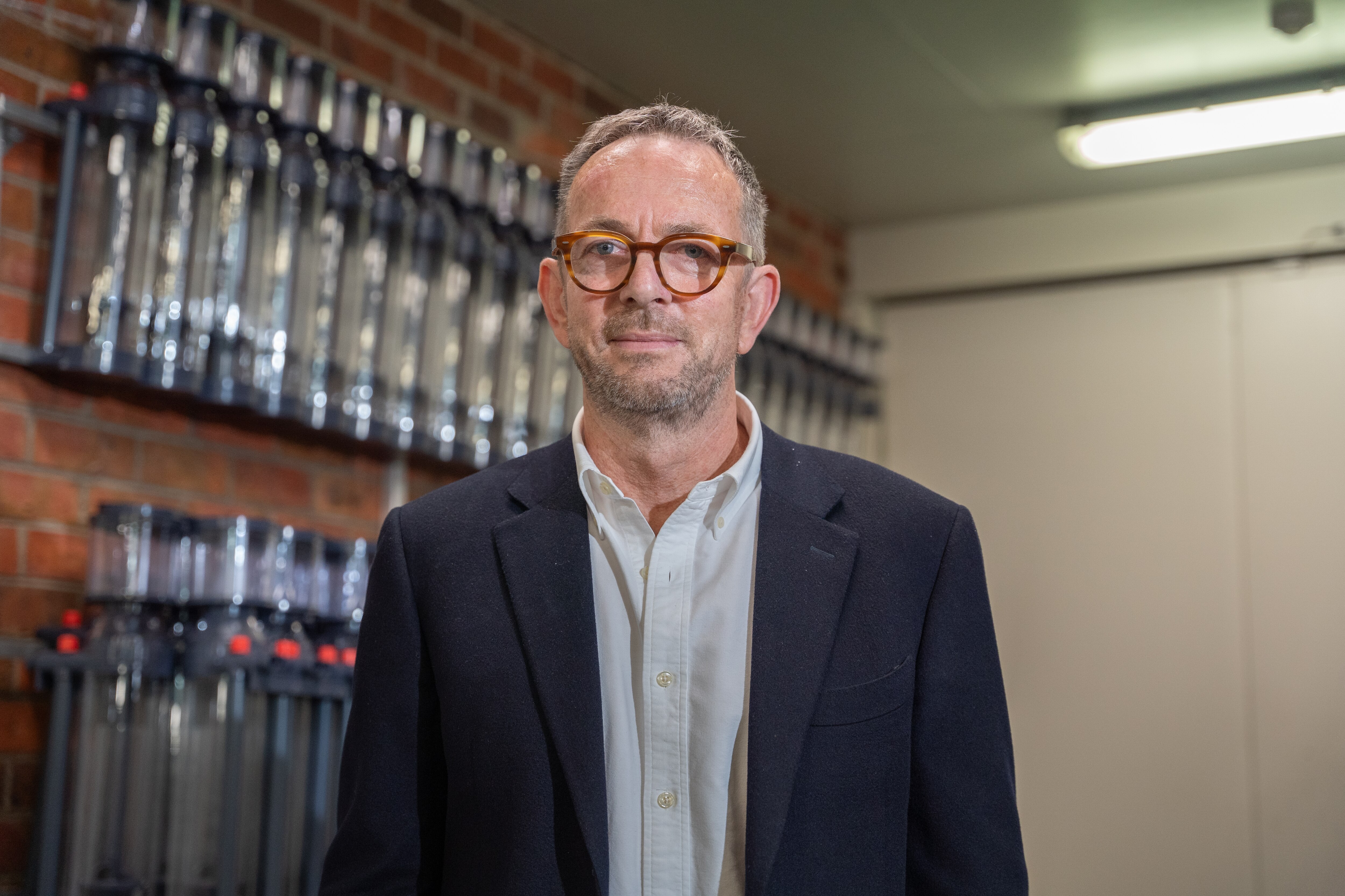 Un hombre de cabello gris con gafas y una chaqueta oscura se encuentra en una habitación con dispositivos pegados a una pared de ladrillos.