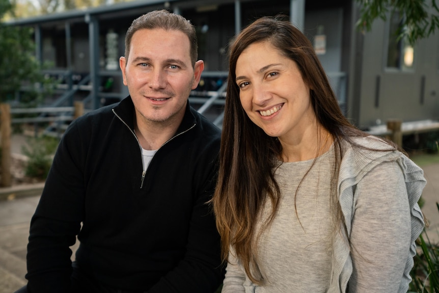 Linda and Mariano Torrisi are sitting next to each other in front of a blue classroom building and smiling.