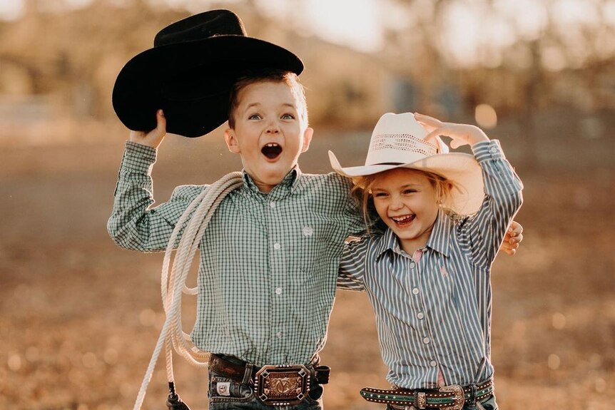 A young boy and girl wearing cowboy gear smile at eachother