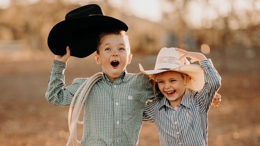 A young boy and girl wearing cowboy gear smile at eachother
