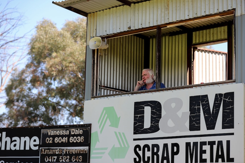Older man with white hair and beard sits in corrugated iron box overlooking football ground 