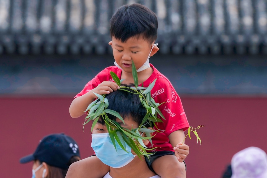 A small boy places a wreath on a masked man's head 