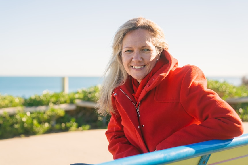 Bourby wears a bright red jacket, sitting on a bench on the coast, smiling at the camera on a sunny day.