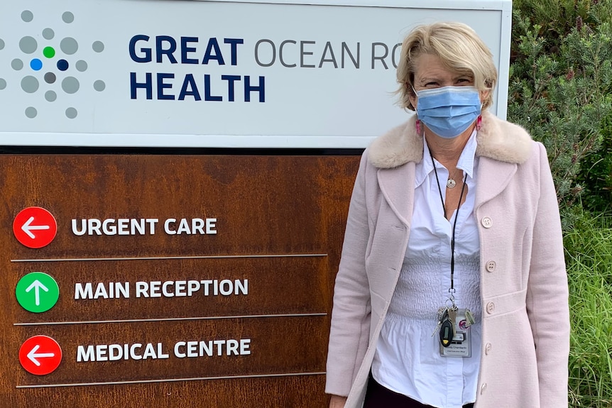 A woman in a long coat and face mask stands in front of a sign that says Great Ocean Road Health