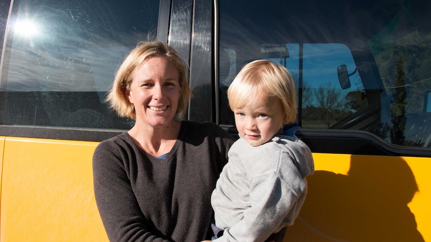 Sam Frost with her son Hugo in her arms standing in front of the yellow school bus she drives