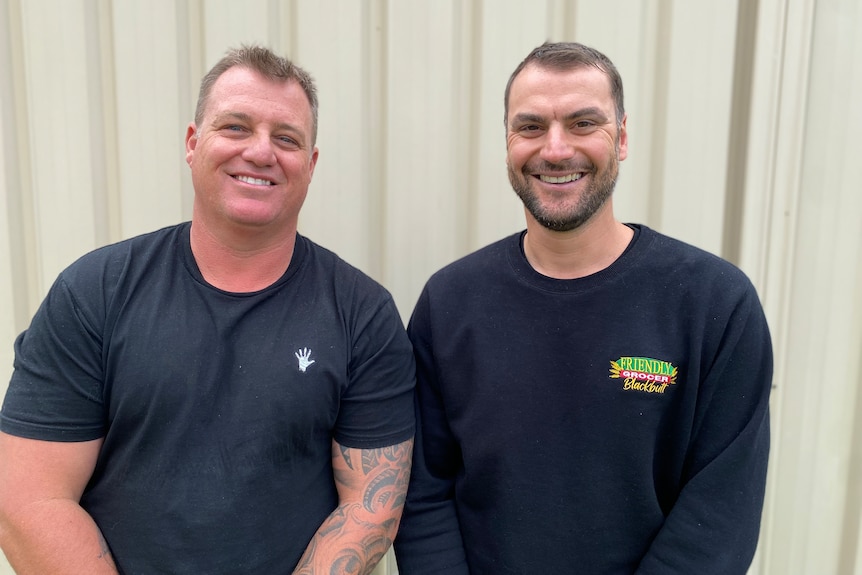 Two smiling men, dressed in black, sitting in front of a metal fence smile at the camera.